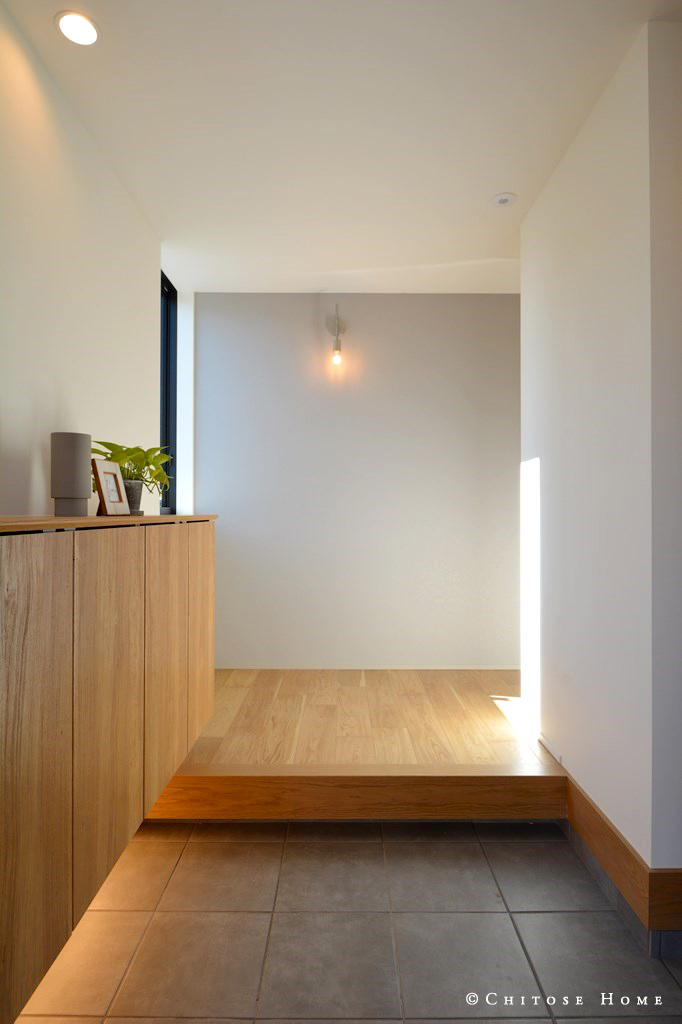 オーク床材の低い彩度に合わせた、照明器具やクロスのコーディネート。こなれ感のある風合いの玄関タイルを間接照明で柔らかく照らす。